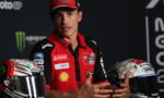 MotoGP rider in a red team polo speaking with hands gesturing, helmets on either side.
