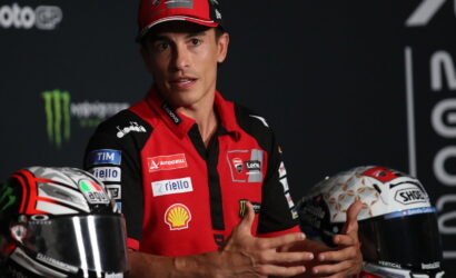 MotoGP rider in a red team polo speaking with hands gesturing, helmets on either side.
