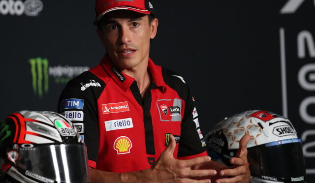 MotoGP rider in a red team polo speaking with hands gesturing, helmets on either side.