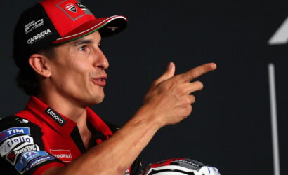 MotoGP rider in red Ducati gear speaks on stage, gesturing with a two-finger point toward the audience