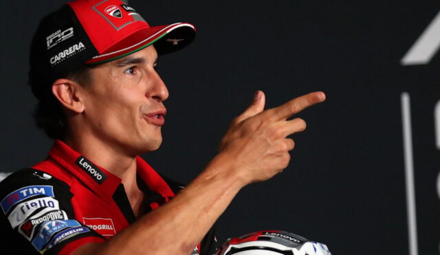 MotoGP rider in red Ducati gear speaks on stage, gesturing with a two-finger point toward the audience