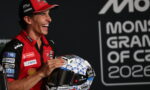 Smiling MotoGP rider in red team gear holds a blue-and-white geometric helmet at a press event.