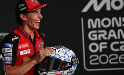 Smiling MotoGP rider in red team gear holds a blue-and-white geometric helmet at a press event.