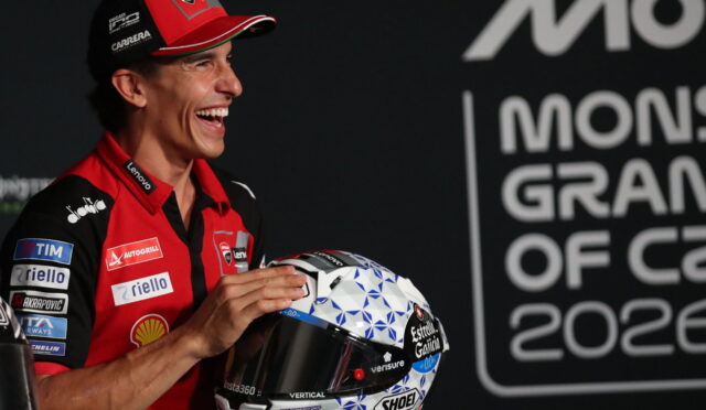 Smiling MotoGP rider in red team gear holds a blue-and-white geometric helmet at a press event.