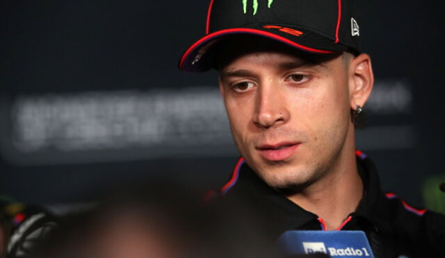 Close-up of a man wearing a black cap with a green Monster logo, speaking at a press conference with a microphone visible in front of him.