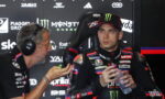 Racing driver in a Monster Energy cap sits in a branded chair, holding a water bottle as a team member with headset talks to him in a sponsor-filled garage backdrop.