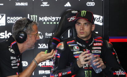 Racing driver in a Monster Energy cap sits in a branded chair, holding a water bottle as a team member with headset talks to him in a sponsor-filled garage backdrop.