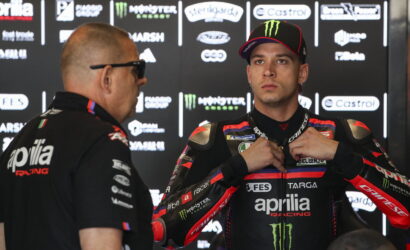 MotoGP racer in a black Aprilia Racing suit adjusts his chest strap during a interview against a sponsor-filled backdrop.