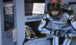 Race motorcycle rider in blue gear and full-face helmet crouches in a motorsport pit area beside monitor screens and charts.
