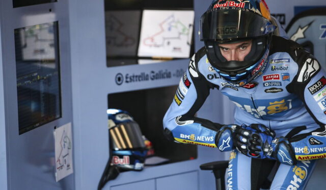 Race motorcycle rider in blue gear and full-face helmet crouches in a motorsport pit area beside monitor screens and charts.