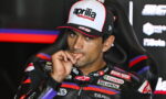 MotoGP rider wearing an Aprilia Racing cap, sitting in a chair with a finger to his lips.