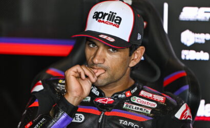 MotoGP rider wearing an Aprilia Racing cap, sitting in a chair with a finger to his lips.