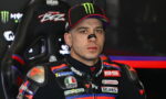 Motorcycle racer in a sponsor-loaded racing suit sitting in a high-backed cockpit seat, wearing a Monster cap.