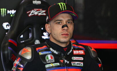 Motorcycle racer in a sponsor-loaded racing suit sitting in a high-backed cockpit seat, wearing a Monster cap.