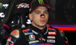 Motorcycle racer in a sponsor-branded suit sits in a racing car cockpit, wearing a Monster cap and nose guard.