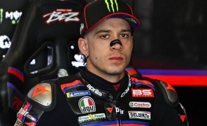 Motorcycle racer in a sponsor-branded suit sits in a racing car cockpit, wearing a Monster cap and nose guard.