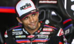 Rider wearing an Aprilia Racing cap sits in a racing seat, smiling slightly, with sponsor logos on his suit.