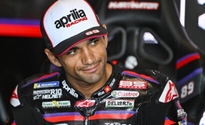 Rider wearing an Aprilia Racing cap sits in a racing seat, smiling slightly, with sponsor logos on his suit.