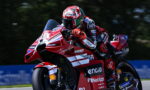 Rider on a red Ducati race bike leaning into a sharp turn, wearing a red-and-white helmet and suit with sponsor logos visible.
