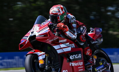 Rider on a red Ducati race bike leaning into a sharp turn, wearing a red-and-white helmet and suit with sponsor logos visible.