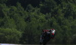 Motorcycle racer in red/black leathers leans into a corner on a race track with dense green trees in the background at high speed.