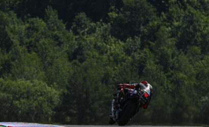 Motorcycle racer in red/black leathers leans into a corner on a race track with dense green trees in the background at high speed.