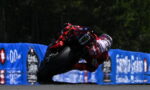 Motorcycle racer in red gear leaning into a turn on a race track with blue barriers in the background.