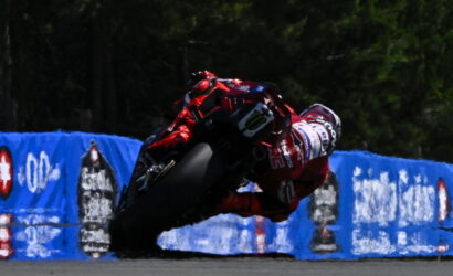 Motorcycle racer in red gear leaning into a turn on a race track with blue barriers in the background.