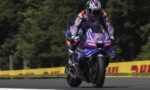 Motorcycle racer in purple racing gear on a purple Yamaha bike leaning into a turn on a race track with yellow safety barriers in the background.
