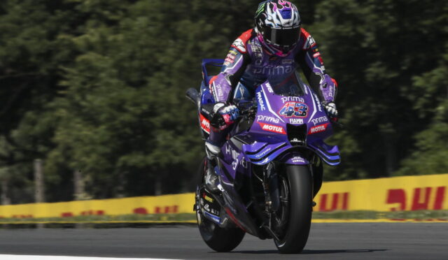 Motorcycle racer in purple racing gear on a purple Yamaha bike leaning into a turn on a race track with yellow safety barriers in the background.