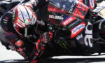 Professional motorcycle racer leaning into a sharp turn, racing on a sponsor-labeled bike with a red helmet in close focus.