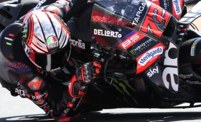 Professional motorcycle racer leaning into a sharp turn, racing on a sponsor-labeled bike with a red helmet in close focus.