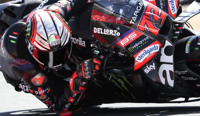 Professional motorcycle racer leaning into a sharp turn, racing on a sponsor-labeled bike with a red helmet in close focus.