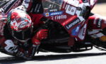 MotoGP rider in red racing suit and Suomy helmet leans into a sharp turn, bike covered in sponsor decals.