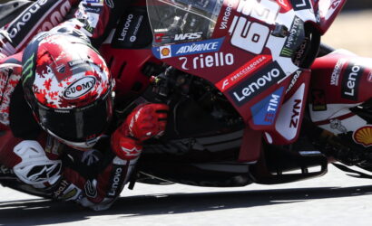 MotoGP rider in red racing suit and Suomy helmet leans into a sharp turn, bike covered in sponsor decals.