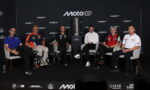 Six men seated in grey chairs on a stage around a tall trophy, with a MotoGP backdrop and sponsor logos behind them.