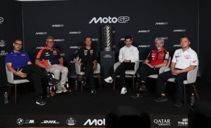 Six men seated in grey chairs on a stage around a tall trophy, with a MotoGP backdrop and sponsor logos behind them.