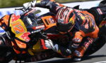 MotoGP rider in orange and black gear leans into a curve on a KTM/Red Bull bike, helmet with red stripes visible in motion on the track.