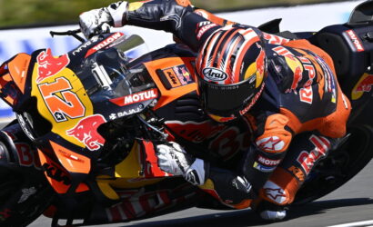 MotoGP rider in orange and black gear leans into a curve on a KTM/Red Bull bike, helmet with red stripes visible in motion on the track.