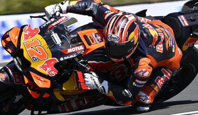 MotoGP rider in orange and black gear leans into a curve on a KTM/Red Bull bike, helmet with red stripes visible in motion on the track.
