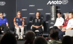 Group of MotoGP riders and team staff seated on a stage for a panel, with a tall trophy on a pedestal to the right and a dark backdrop featuring MotoGP logos.