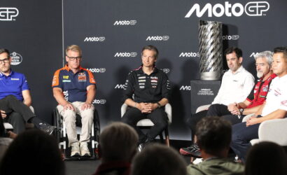 Group of MotoGP riders and team staff seated on a stage for a panel, with a tall trophy on a pedestal to the right and a dark backdrop featuring MotoGP logos.