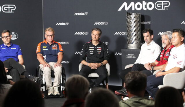 Group of MotoGP riders and team staff seated on a stage for a panel, with a tall trophy on a pedestal to the right and a dark backdrop featuring MotoGP logos.