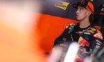 Moto racer in orange-and-black racing suit sits in a pit area, looking upward with sponsor banners in the background (Interwetten, Red Bull).