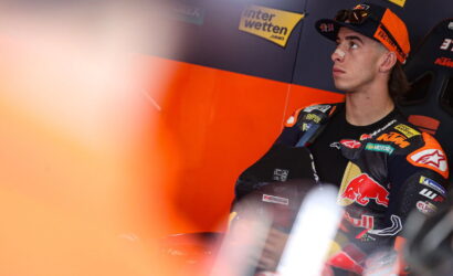 Moto racer in orange-and-black racing suit sits in a pit area, looking upward with sponsor banners in the background (Interwetten, Red Bull).
