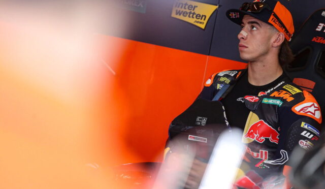 Moto racer in orange-and-black racing suit sits in a pit area, looking upward with sponsor banners in the background (Interwetten, Red Bull).