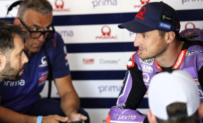 Motorcycle racer in purple gear talks with team member in a pit area backdrop of sponsor banners