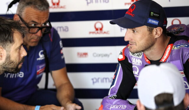 Motorcycle racer in purple gear talks with team member in a pit area backdrop of sponsor banners