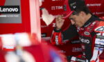 MotoGP rider in a red Lenovo racing suit adjusts his cap in the pit area.