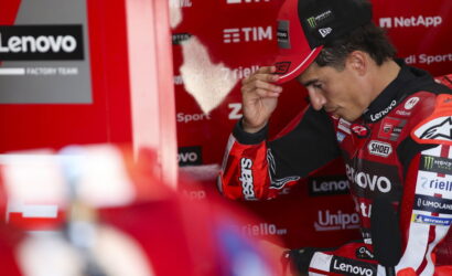 MotoGP rider in a red Lenovo racing suit adjusts his cap in the pit area.